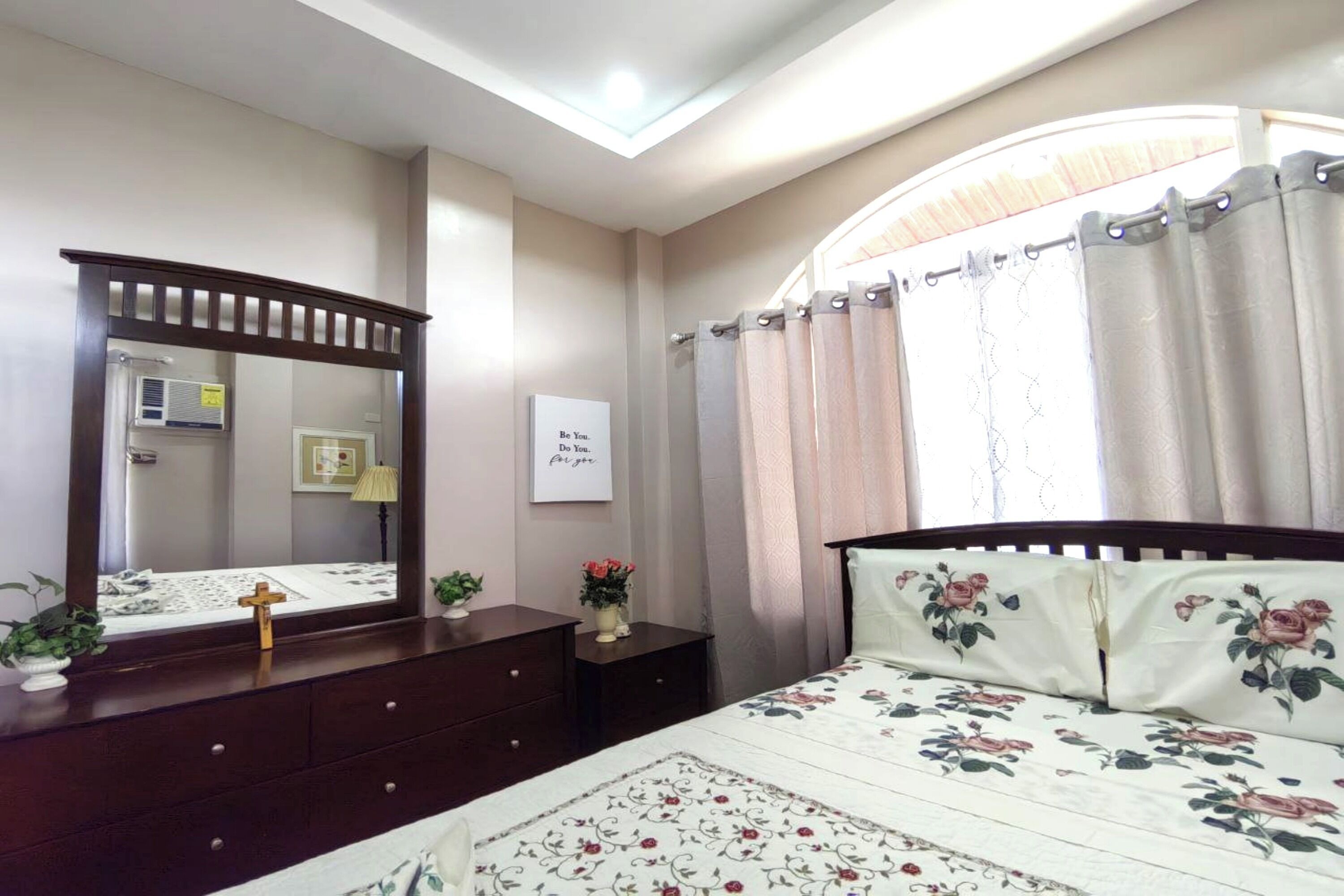 Um quarto com uma cama, penteadeira, espelho, janela com cortinas e ar condicionado fixado na parede.