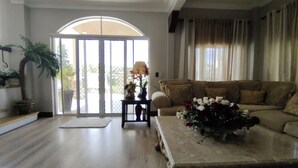 Ein Wohnzimmer mit einer Couch, einem Couchtisch mit floraler Dekoration, einer Topfpflanze und einer Glastür, die zu einem Außenbereich führt.