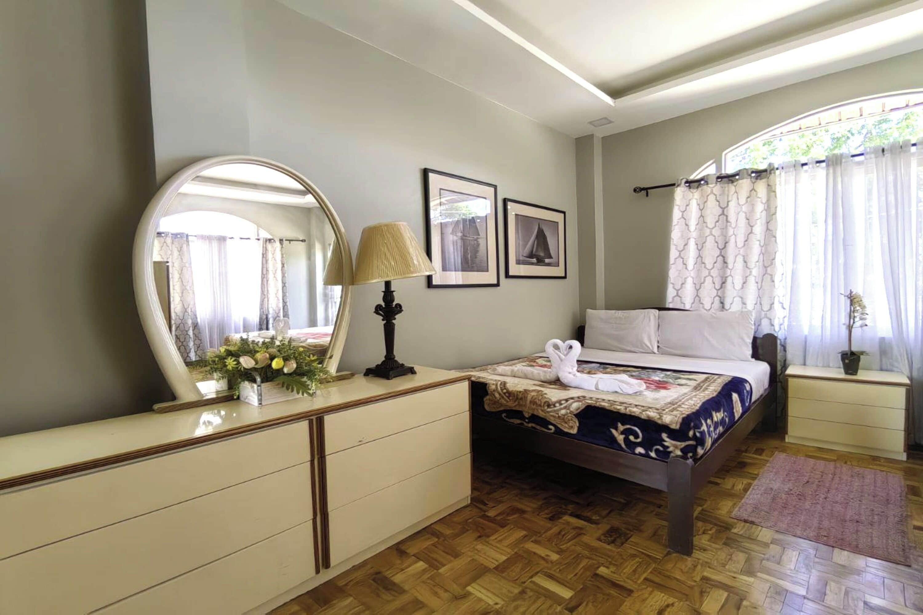 A bedroom with a wooden floor, a bed with a decorative headboard, a bedside table with a lamp, a dresser, and a large mirror.