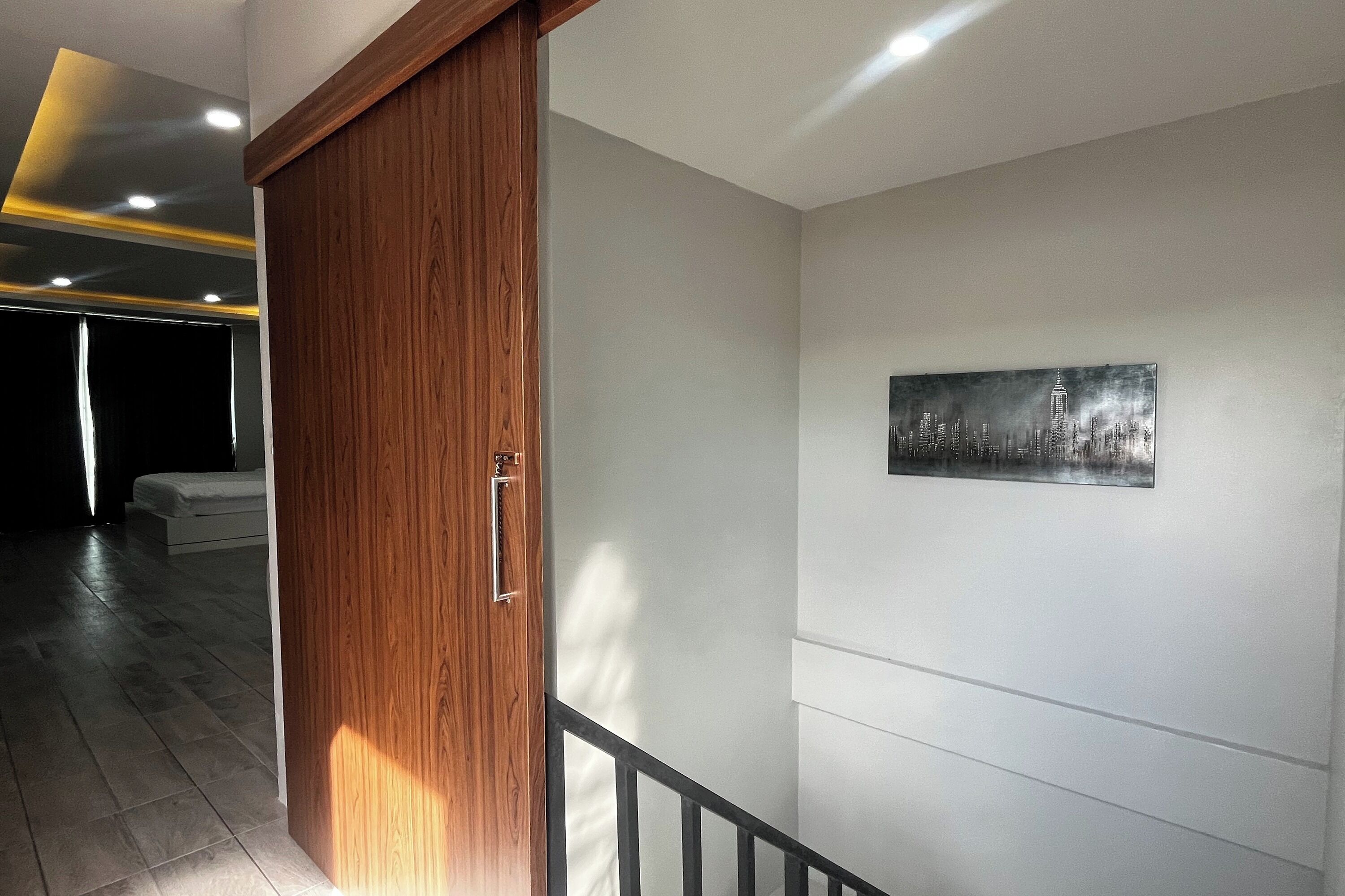 Un interior moderno con una puerta de madera, una pared blanca con una obra de arte de un paisaje urbano y una escalera con barandilla negra.