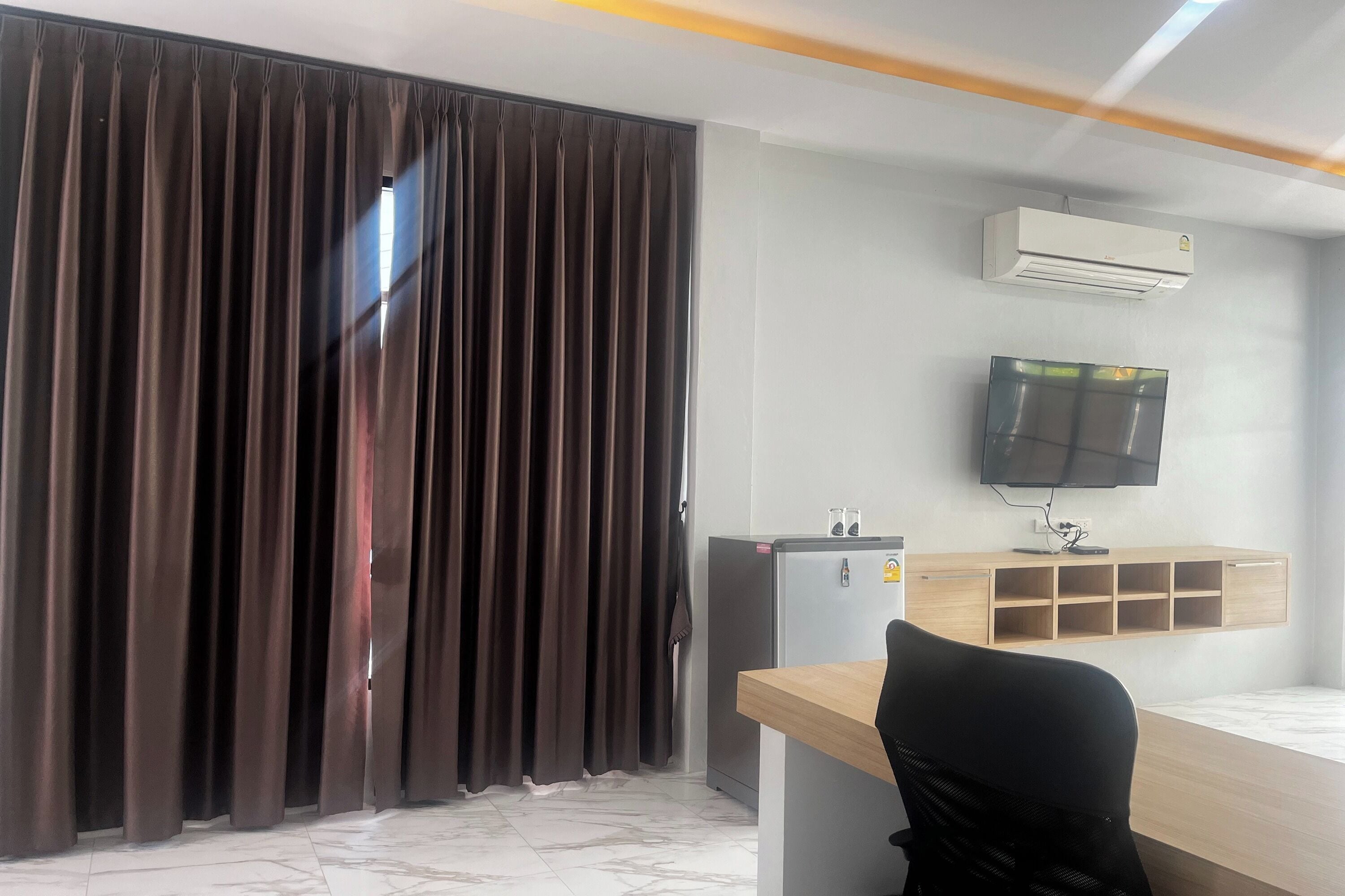 A hotel room with a large window, a desk with a chair, an air conditioner, a television, and a minibar.
