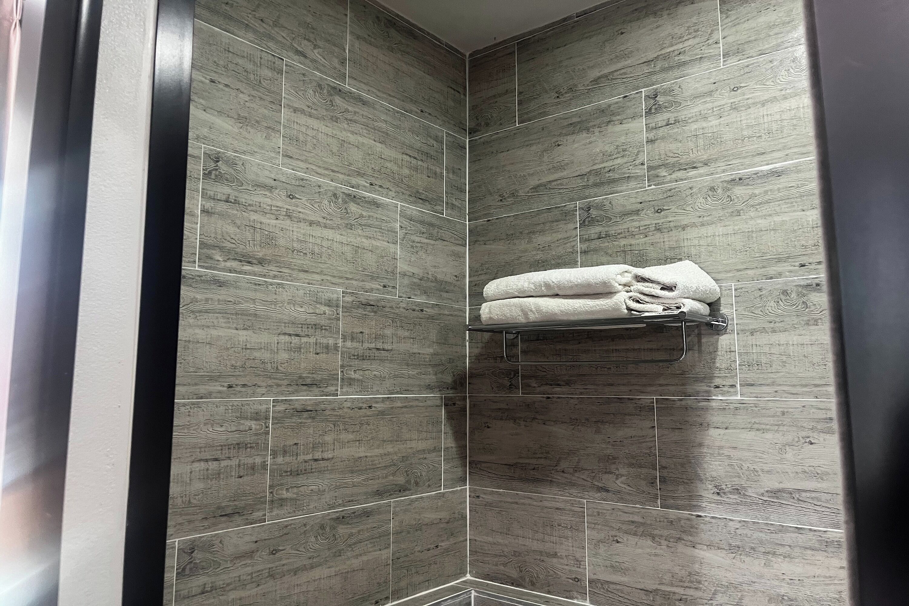 Ducha en esquina con azulejos de madera gris y un perchero con toallas dobladas.