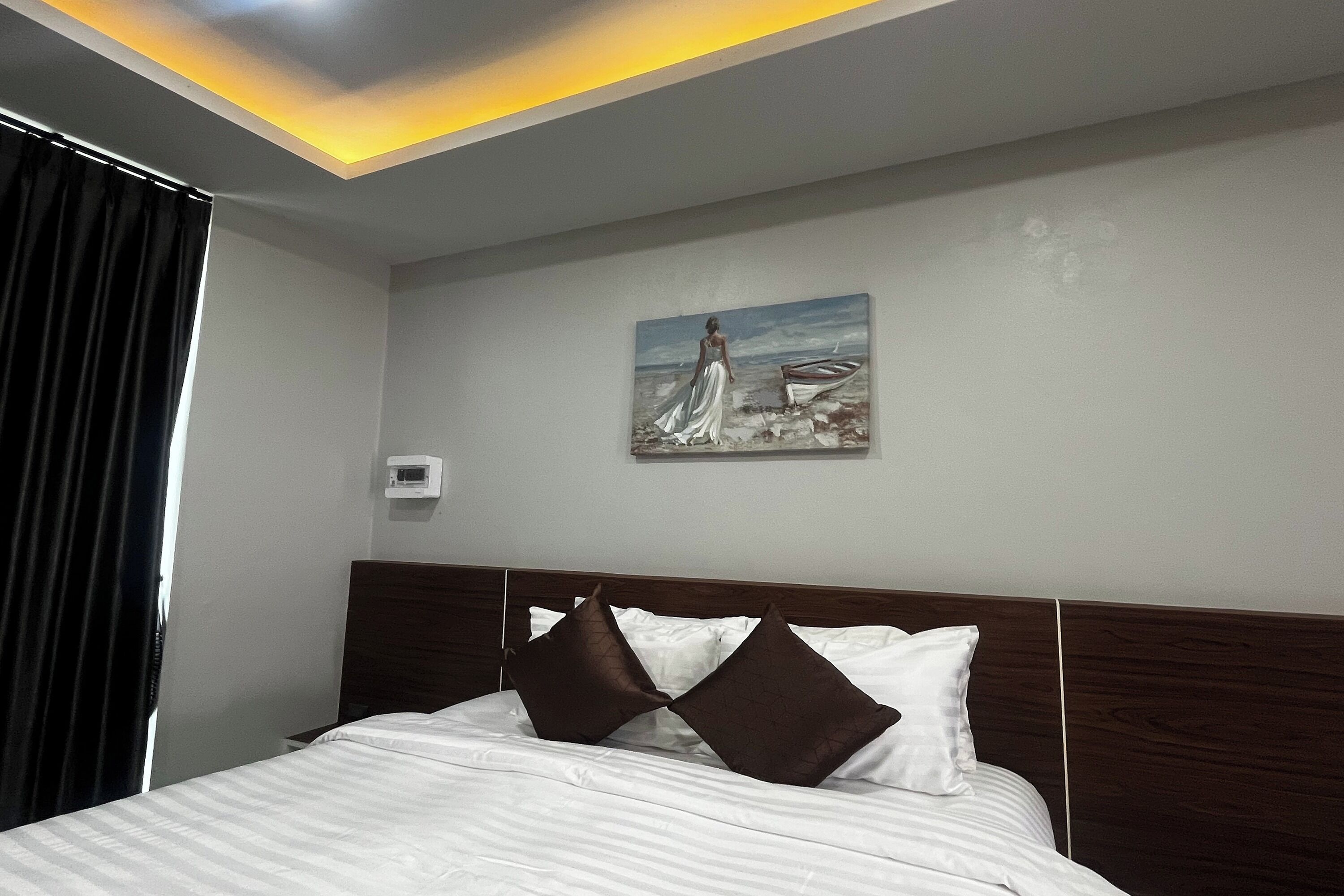 Habitación de hotel con una cama, dos almohadas, un cuadro de una mujer junto al mar y una lámpara en el techo.