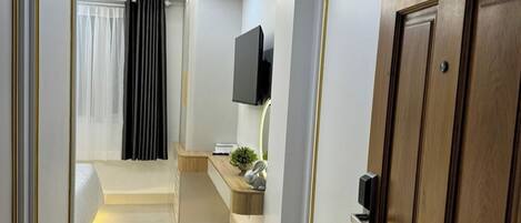 Une chambre d’hôtel moderne avec une porte en bois, un téléviseur à écran plat fixé au mur, une table de chevet avec une lampe et une petite plante en pot.