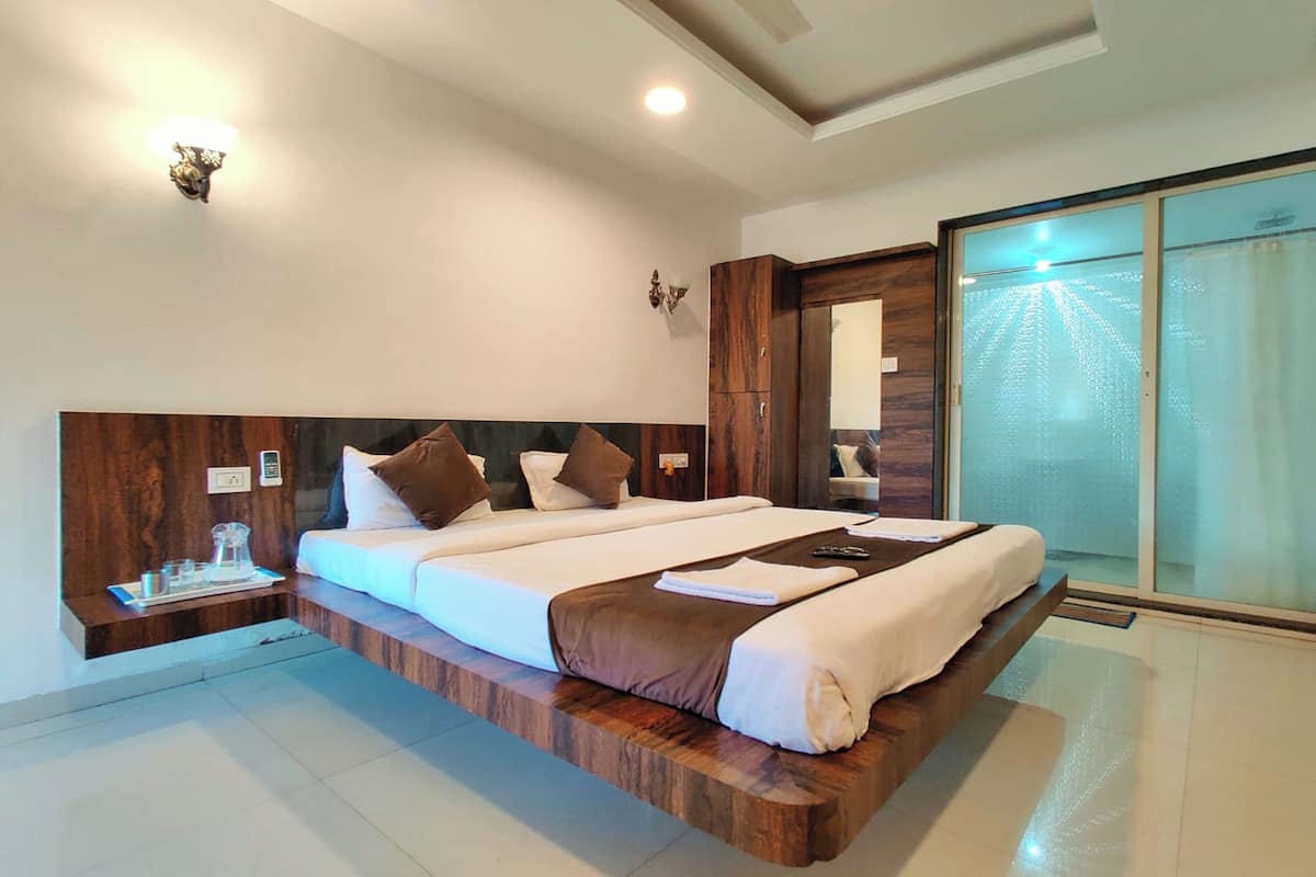 A modern hotel room with a large bed, wooden headboard, bedside tables with glassware, and a sliding glass door leading to a balcony.