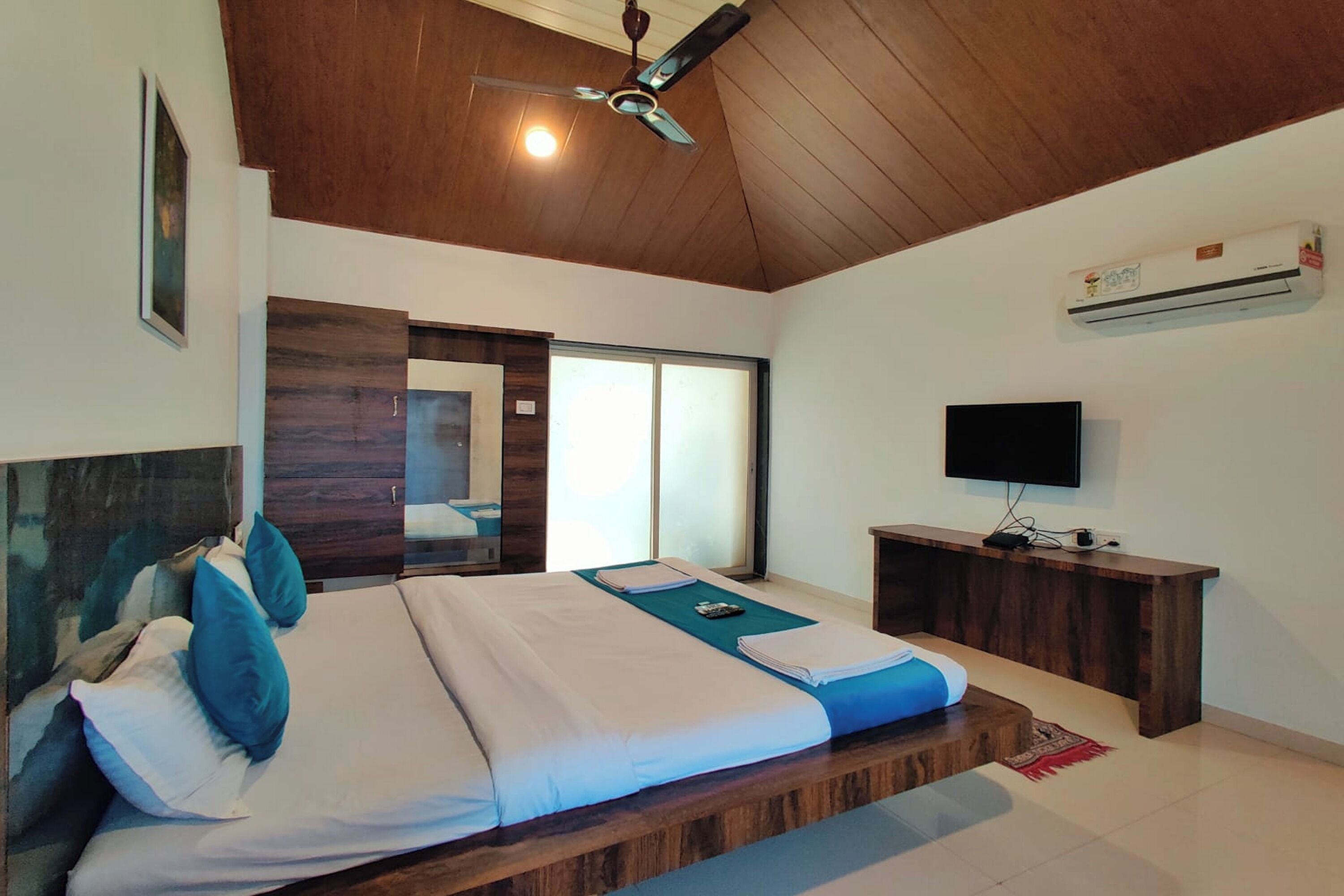 Una camera da letto con soffitto in legno, un letto con biancheria azzurro e bianca, una TV su un mobiletto in legno e un condizionatore d'aria a parete.