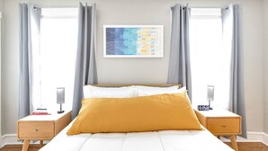 Ein Schlafzimmer mit einem Bett, zwei Nachttischen, einem Wandbild und zwei mit Vorhängen versehenen Fenstern.