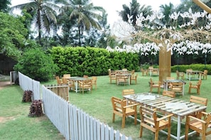 An outdoor dining area with multiple tables and chairs, surrounded by lush greenery and palm trees. - Seed Resort (Shrirangapattana)