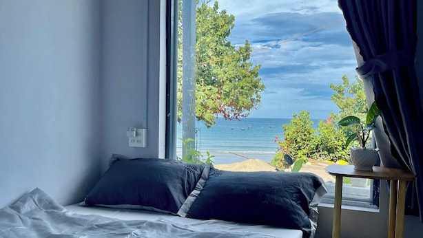 Une chambre avec une grande fenêtre, un lit recouvert d’une literie sombre, une petite table en bois et une vue sur la mer et la verdure.