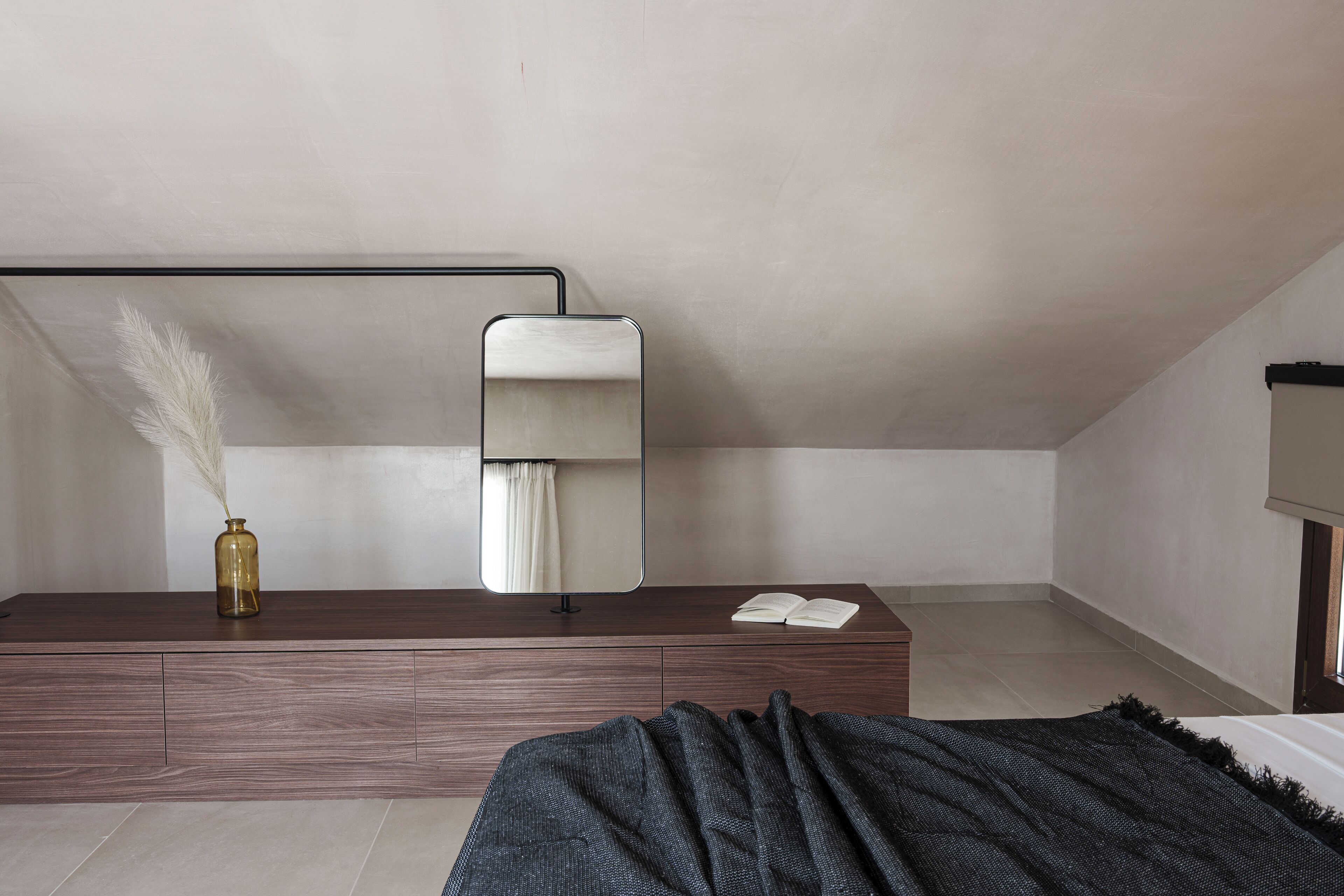 A minimalist bedroom with a wooden dresser, a mirror, and a bedside table with a book.
