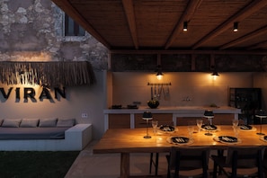 Un comedor rústico con una mesa de madera dispuesta para seis comensales, un banco empotrado y una cocina al fondo.