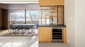 A modern kitchen with a built-in wine cooler, a dining area with a table and chairs, and a view of trees through large windows. - Villa Lily (Hakuba)