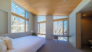 A bedroom with a large bed, a desk, and a chair, with large windows offering a view of trees. - Villa Lily (Hakuba)