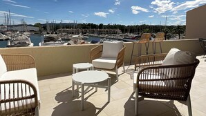 Eine Terrasse mit Korbmöbeln, einem weißen Tisch und Blick auf einen Yachthafen mit Booten.
