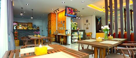 A modern restaurant with wooden tables and chairs, a kitchen area, and a TV mounted on the wall.