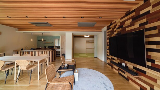 Een moderne woonkamer met een houten plafond, een grote flatscreen televisie aan een muur bevestigd, een eethoek met rieten stoelen, en een keuken op de achtergrond.