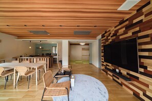 A modern living room with a wooden ceiling, a large flat-screen TV mounted on a wall, a dining area with wicker chairs, and a kitchen in the background. - Yamakitano Akari (Ukiha)