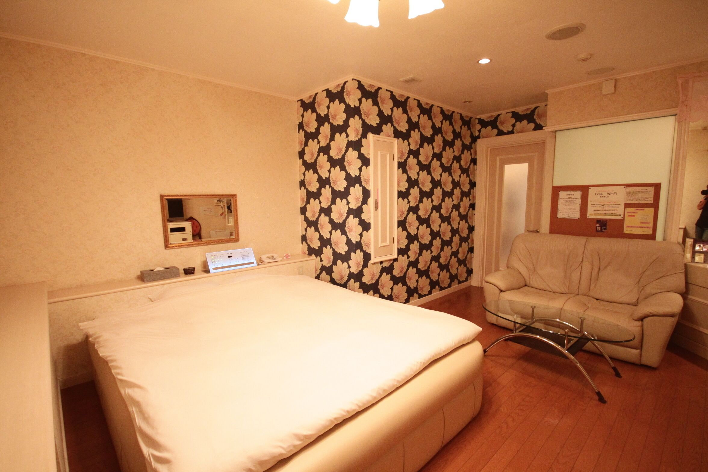 A hotel room with a bed, a chair, a desk with a computer, and a bulletin board with notices.