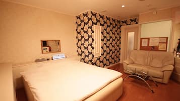 A hotel room with a bed, a chair, a desk with a computer, and a bulletin board with notices.