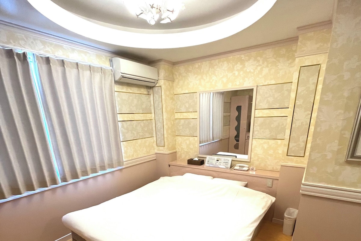 A hotel room with a large bed, a bedside table with a lamp, a mirror, a window with curtains, and an air conditioning unit.