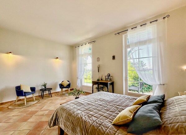 A bedroom with a large bed, a chair, a small table, and a window with curtains.