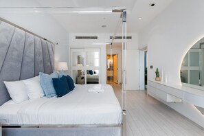 A modern bedroom with a large bed, a bedside table with a lamp, a mirror, and a view of the hallway.
