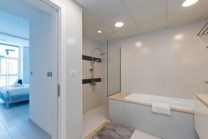 A modern bathroom with a glass shower enclosure, a white bathtub, and a visible bed in the adjacent room.