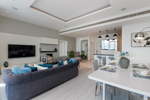 Ein modernes Wohnzimmer mit grauem Sofa, einem Flachbildfernseher und einem Essbereich mit weißem Tisch und Stühlen.