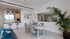 Un espace repas moderne avec une table blanche, des chaises grises et des tabourets de bar. On y trouve une grande plante en pot et un canapé avec des coussins bleus.