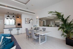 Een moderne eetruimte met een witte tafel, grijze stoelen en barkrukken. Er staat een grote potplant en een bank met blauwe kussens.