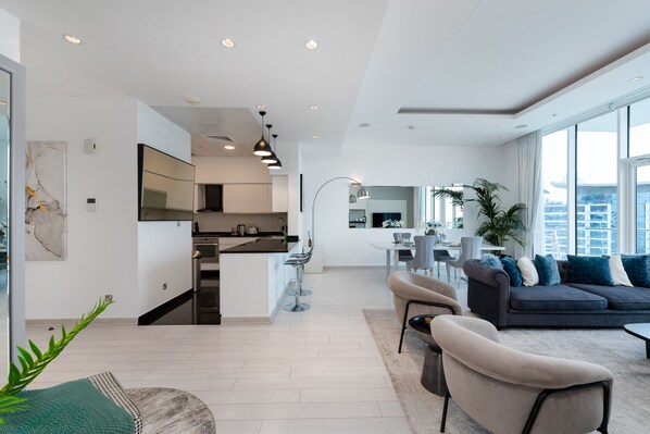 Ein modernes Wohnzimmer mit einem dunkelgrauen Sofa, beigen Sesseln und einem Essbereich mit einem weißen Tisch und Stühlen.