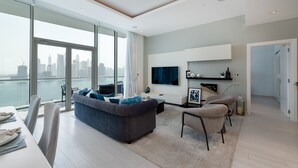 Un salon moderne doté d’une grande fenêtre donnant sur un paysage urbain, d’un canapé gris, d’un coin repas et d’un téléviseur à écran plat fixé au mur.