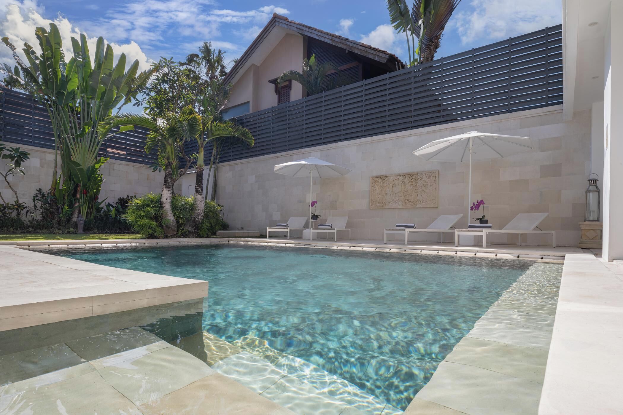 A clear swimming pool with lounge chairs and umbrellas, surrounded by tropical plants and a modern building.