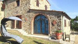 A stone building with a large arched window, a small porch with a chair, and a potted plant. - Villa in Tuscany with Pool & Olive Groves (Castelnuovo di Val di Cecina (PI))