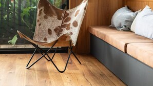 A room with a wooden floor, a cushioned bench with pillows, a patterned chair, and a large window with a view of greenery. - Loft Summary:<br>Within (Bangalow)