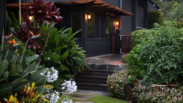 A modern house with a dark exterior, a covered porch, and a well-maintained garden with various plants and flowers. - Loft Summary:<br>Within (Bangalow)