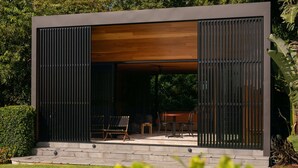A modern outdoor seating area with a wooden ceiling and black metal shutters, surrounded by lush greenery. - Three One Bedroom Cabins (Bangalow)