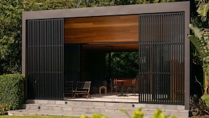 A modern outdoor seating area with a wooden ceiling and black metal shutters, surrounded by lush greenery.