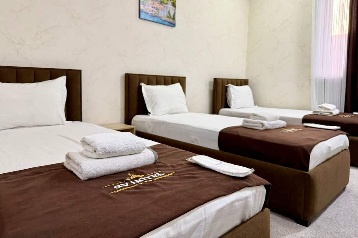 A hotel room with three beds, each with a brown headboard and white bedding. There is a painting on the wall above the beds.