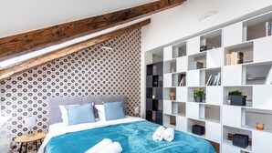Een moderne slaapkamer met een schuine plafond, behang met een patroon, een groot bed met een blauwe deken, en een witte inbouwkast met diverse spullen.