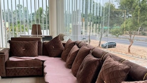 A modern living room with a large sectional sofa, a ceiling fan, and a view of the street. - Hotel Ramsons International  (Hunsur)