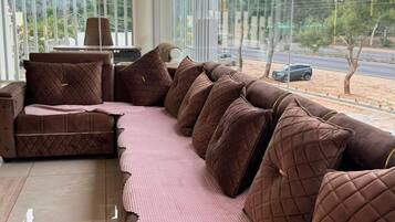 Un área de descanso moderna con un sofá grande y sillones, un ventilador de techo y vistas a la calle a través de amplios ventanales.