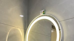 A bathroom with a white sink, a round mirror with a light, and a small green box on the wall. - Hotel Ramsons International  (Hunsur)