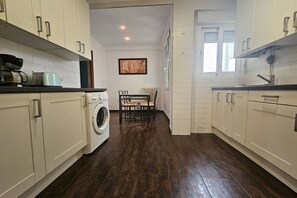 A kitchen with white cabinets, a washing machine, a dining table with chairs, and a framed picture on the wall.
