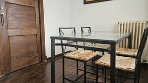 A dining area with a glass-top table, four chairs, a wooden door, and a radiator. - 10a05 Apartamento 2 dormitorios vistas al parque (Pravia)