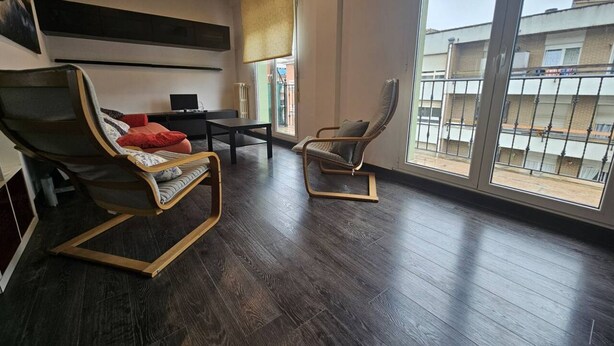 A spacious room with a dark wooden floor, a large window with a view of buildings, a sofa, a chair, and a small table. - 10a05 Apartamento 2 dormitorios vistas al parque (Pravia)