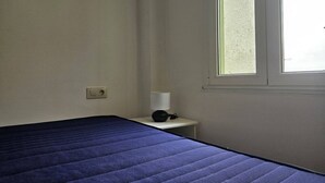 Ein Zimmer mit einem Bett, einem Nachttisch mit Lampe, einem Fenster und einer Steckdose.