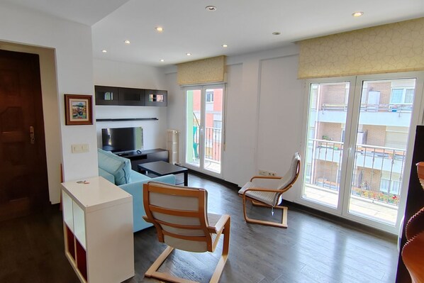 A modern living room with a sofa, two chairs, a TV, and a kitchen area.