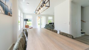 A spacious dining area with a long wooden table and cushioned chairs, a modern pendant light fixture, and a view of the outdoors through large windows. - Holiday Home by Lake Beach Simonshaven (Simonshaven)