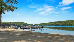 A lakeside dock with boats, a sandy beach, and forested hills in the background.